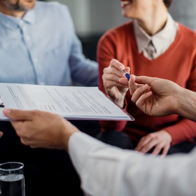 Close-up of woman signing an agreement with insurance agent while being on a meeting with her husband.
